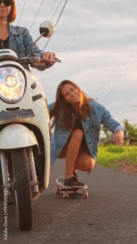 Two young women enjoying an evening ride in Bali, one driving a scooter while the other skateboards along a rural road. Lush rice fields and tall palm trees frame the tropical landscape as the warm