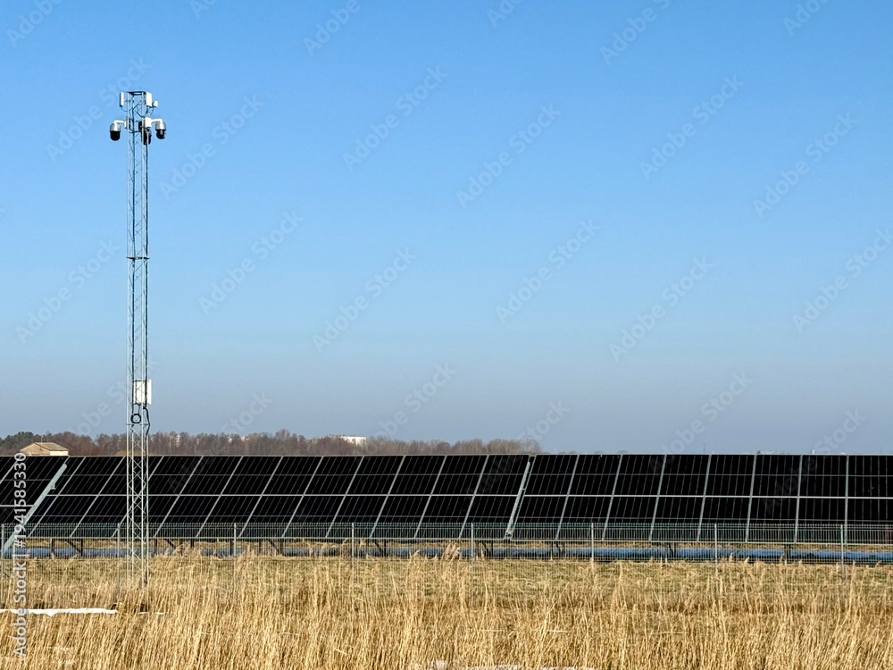 Fototapeta premium Solar panel installation in an open field with dry grass and a tall light pole under clear sky.