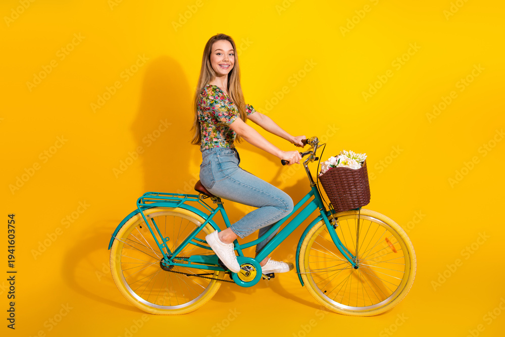Naklejka premium Young woman rides a turquoise bicycle with a wicker basket full of flowers against a bright yellow background