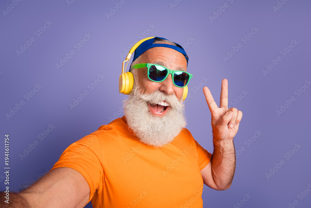 Naklejka premium Happy senior man with yellow headphones and green sunglasses, orange tee, peace sign, and playful energy against a purple background