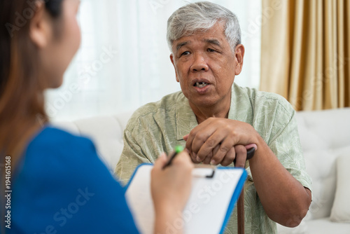 Professional Asian nurse taking notes on clipboard while consulting senior man at home. Geriatric care, home healthcare service, and medical support for elderly people in living room concept