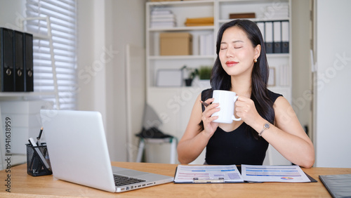 Businesswoman enjoying a coffee break at her desk, holding a mug with eyes closed while working with financial reports and a laptop in a bright office.
