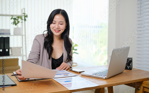 Young entrepreneur reviewing business data and charts on paperwork, sitting at a desk with a laptop.