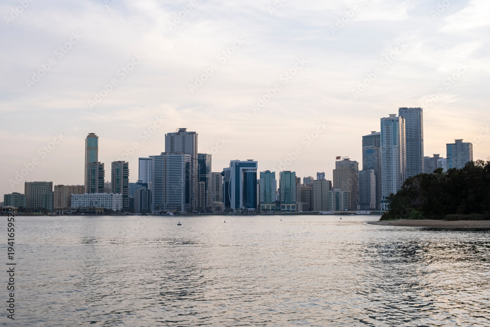 Obraz premium 12.12.2025 Sharjah, UAE. Panoramic view of Sharjah Corniche at sunset, featuring Khalid Lagoon water, illuminated skyscrapers, modern city skyline, and silhouette of date palm trees in UAE
