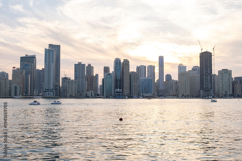 Obraz premium 12.12.2025 Sharjah, UAE. Panoramic view of Sharjah Corniche at sunset, featuring Khalid Lagoon water, illuminated skyscrapers, modern city skyline, and silhouette of date palm trees in UAE
