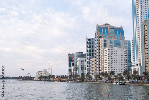 12.12.2025 Sharjah, UAE. Panoramic view of Sharjah Corniche at sunset, featuring Khalid Lagoon water, illuminated skyscrapers, modern city skyline, and silhouette of date palm trees in UAE
