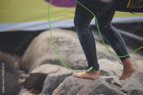 Close-up of a surfer’s ankle secured with a safety leash while stepping toward the ocean, capturing the anticipation of entering the waves. The image highlights the connection between surfer and board