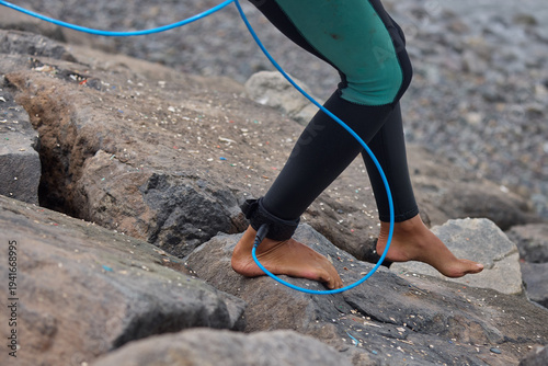 Close-up of a surfer’s ankle secured with a safety leash while stepping toward the ocean, capturing the anticipation of entering the waves. The image highlights the connection between surfer and board