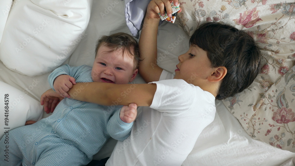 Fototapeta premium Baby annoyed as older brother rests arm on chest, baby reacts by pinching sibling’s arm, playful sibling quarrel, childhood mischief, authentic family dynamics, funny moment