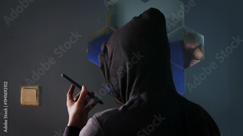 Anonymous spy person or hacker with black hoodie talking on the phone indoors. Standing near a mirror in the room. Mysterious man making phone call conversation with his back to camera. 