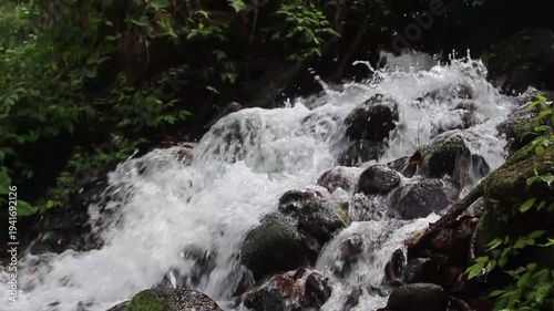 Dynamic Forest Waterfall: Rushing Water Cascading Over Mossy Rocks Amidst Lush Green Foliage