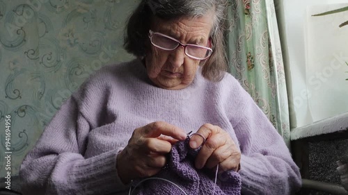 A close-up of an elderly woman diligently knitting a purple garment, showcasing her focus and skill in a quiet home setting.