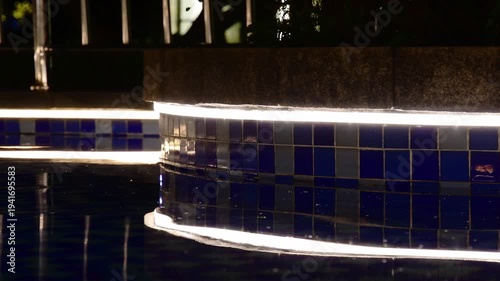 Illuminated swimming pool edge at night with sparkling blue mosaic tiles reflected in tranquil dark water. Modern luxury ambiance for relaxation