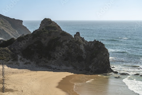 Rocky coastline and sandy beach along the Costa Vicentina, Algarve, Portugal. A scenic view of rugged cliffs meeting golden sand and calm ocean waves on a sunny day in southern Portugal.