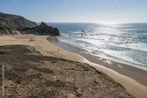 Coastal landscape of Costa Vicentina, Algarve, Portugal with sandy beach and rocky cliffs. Aerial perspective of a sandy beach bordered by rugged cliffs under clear blue sky, with surfers in the ocean