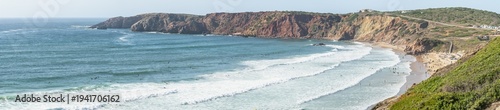 Panoramic view of Praia do Amado at Costa Vicentina coastline with surfers, cliffs, and ocean waves in Algarve, Portugal. Coastal landscape showing waves breaking on sandy beach, rocky cliffs, surfers