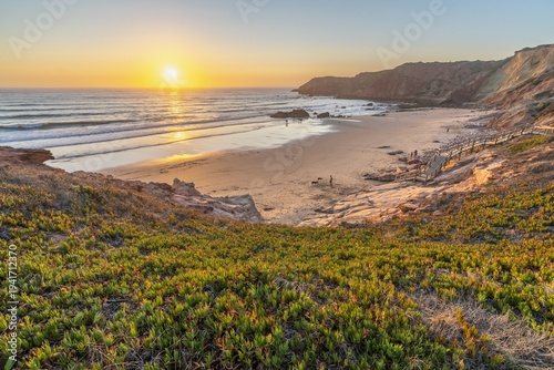 Sunset over  Praia do Amado at Costa Vicentina beach in Algarve, Portugal. Golden sunset illuminating a serene beach with waves, dunes, and rugged cliffs along the Costa Vicentina coastline.