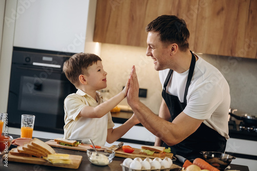 Wallpaper Mural Father and young son sharing a high-five in a modern kitchen, surrounded by breakfast ingredients including bread, eggs, and vegetables on a wooden table Torontodigital.ca