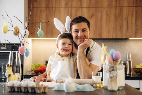 Wallpaper Mural Father and son wearing bunny ears smile together in a modern kitchen, surrounded by Easter decorations, colorful eggs, and baking supplies on the countertop Torontodigital.ca