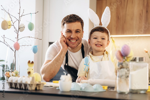 Wallpaper Mural Father and son wearing bunny ears smile while decorating Easter eggs at kitchen counter with colorful decorations and festive elements in the background Torontodigital.ca