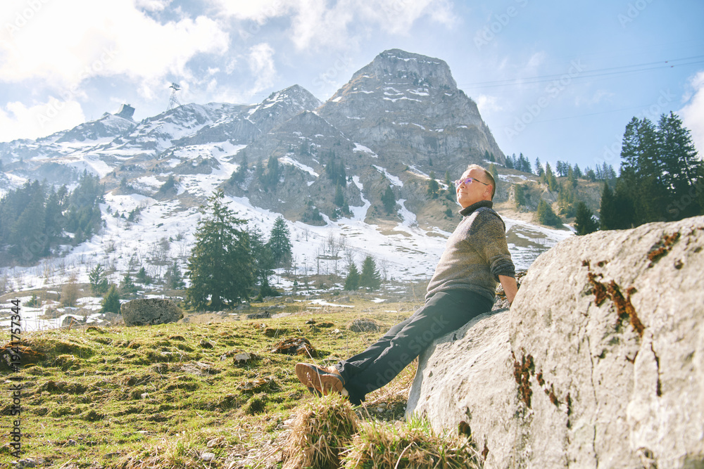 Fototapeta premium Senior man relaxing on rock in Swiss Alps near Gruyere, enjoying sunny mountain landscape with snow peaks, peaceful travel lifestyle and nature freedom