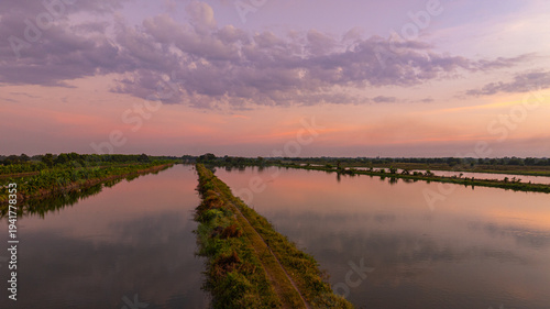 A stunning golden sunset reflecting on a calm lake surrounded by rural greenery and tree silhouettes. The dramatic clouds and warm evening light create a peaceful natural landscape perfect for travel