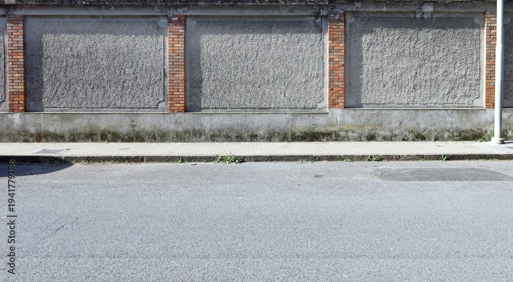 Fototapeta premium Grunge rough concrete wall with rough brick columns. Tiled sidewalk and street in front. Urban background for copy space.