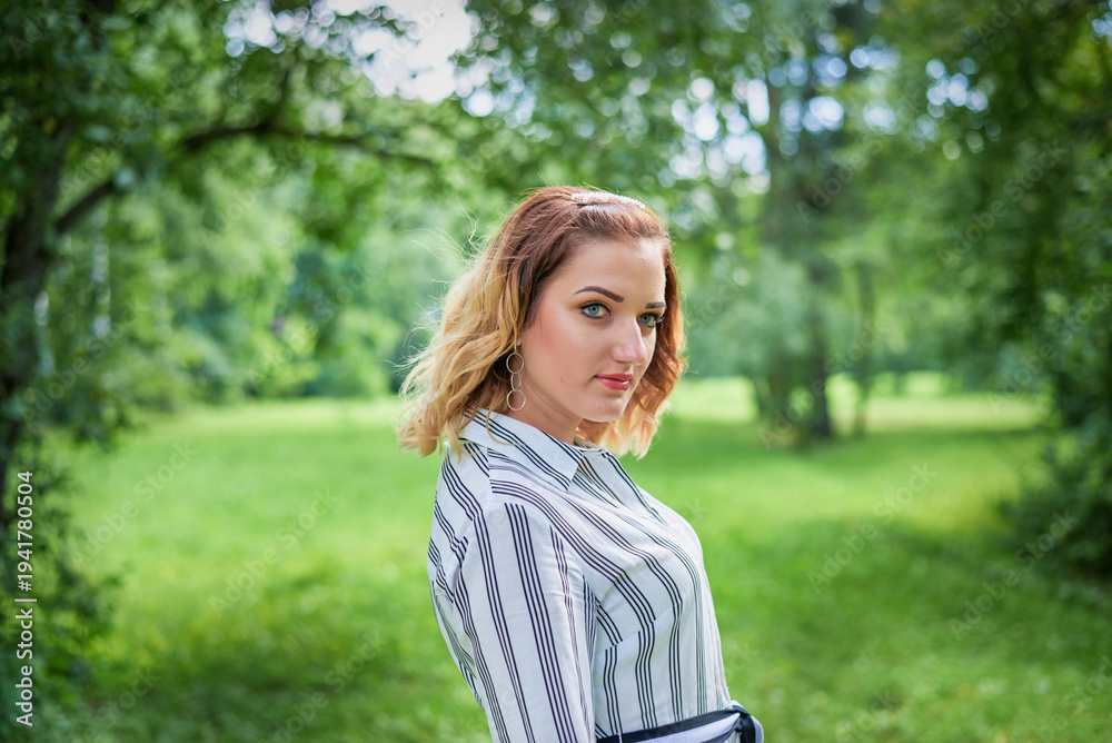 Obraz premium A woman in a white dress stands in a sunlit forest clearing on a summer day. In front of her is lush green grass, and behind her are tall, slender trees.