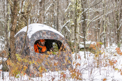 Male hunter in a hunting blind wearing safety orange camouflage and holding a rifle in the woods with fresh snow looking for deer. Killington, Vermont.
