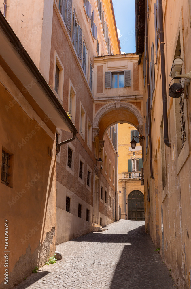 Fototapeta premium Quiet narrow street in the historic center of Rome with stone pavement and arch connecting old buildings.
