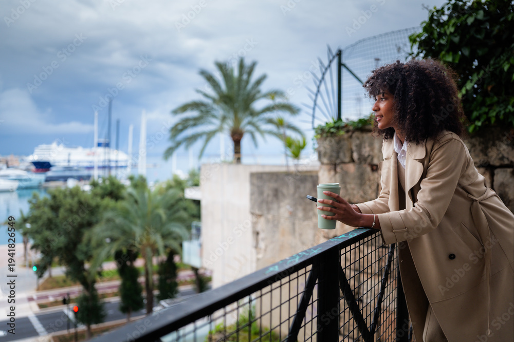 Fototapeta premium Young black woman leans on balcony railing with reusable coffee cup and smartphone, gazing over a harbor with boats and palm trees, enjoying a peaceful urban moment