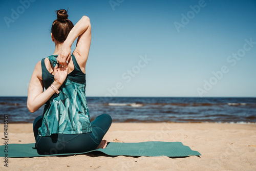 Woman stretching shoulders and back while sitting on yoga mat at sandy beach. Ocean background and clear sky create relaxing atmosphere focused on flexibility, posture and wellness.