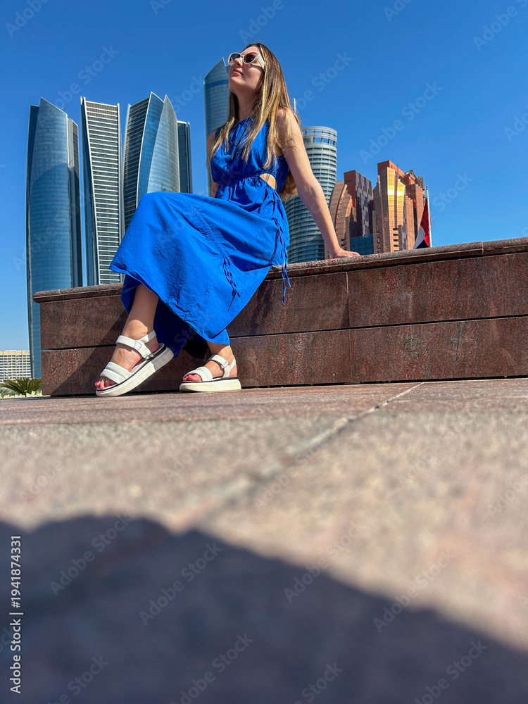Fototapeta premium Fashionable woman in blue dress sitting on terrace with modern skyscraper skyline, sunny urban travel lifestyle