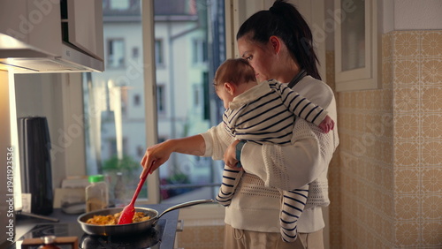 Εκτύπωση καμβά Mother holding baby while cooking in kitchen, stirring food in frying pan, multi