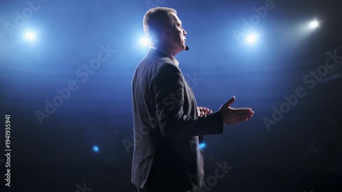 Senior male motivational speaker passionately gestures while delivering an inspiring speech on stage, wearing a headset microphone. Vibrant stage lights illuminate the scene, creating a dynamic and