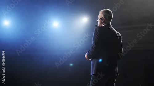 Senior male motivational speaker passionately gestures while delivering an inspiring speech on stage, wearing a headset microphone. Vibrant stage lights illuminate the scene, creating a dynamic and