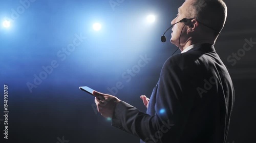 Businessman confidently presenting on stage at a conference, using a digital tablet and wearing a headset microphone. Bright spotlights and atmospheric stage fog create a dramatic setting as he
