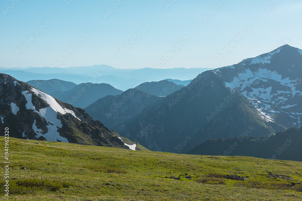 Fototapeta premium Scenic view from sunlit grassy meadow to abyss of alpine valley between big rocky mountains with snows and hilly layers of ridge silhouettes far away under clear blue sky in bright sun in sunny day.