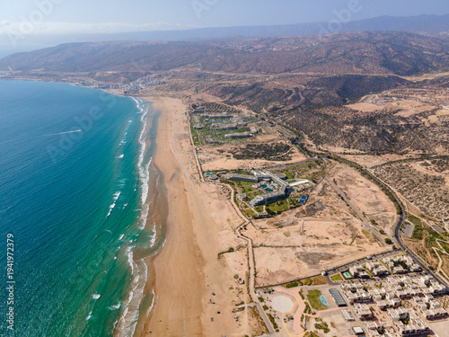 Aerial drone view of the beautiful Taghazout Bay coastline featuring luxury resorts, sandy beaches, lush green golf courses, and the Atlantic Ocean in Agadir, Morocco.