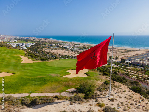 Aerial drone view of the beautiful Taghazout Bay coastline featuring luxury resorts, sandy beaches, lush green golf courses, and the Atlantic Ocean in Agadir, Morocco.