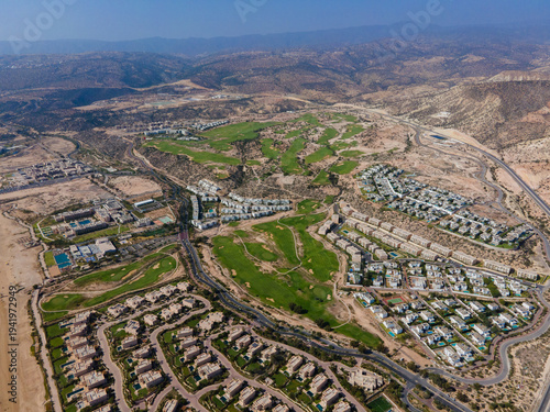 Aerial drone view of the beautiful Taghazout Bay coastline featuring luxury resorts, sandy beaches, lush green golf courses, and the Atlantic Ocean in Agadir, Morocco.
