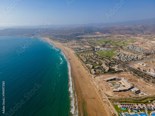 Aerial drone view of the beautiful Taghazout Bay coastline featuring luxury resorts, sandy beaches, lush green golf courses, and the Atlantic Ocean in Agadir, Morocco.