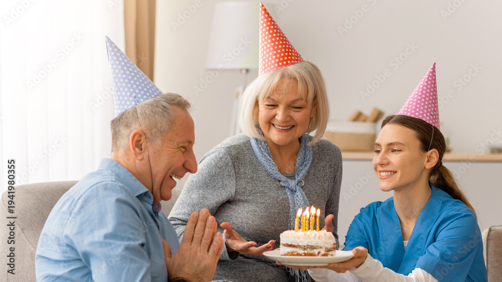 Obraz premium A man smiles and claps as he receives a birthday cake from a woman wearing blue scrubs. Another woman with gray hair smiles beside him. They celebrate together in a warm living room.