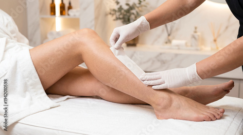 Beautician wearing white gloves removing hair from a client's leg using a wax strip in a bright beauty salon