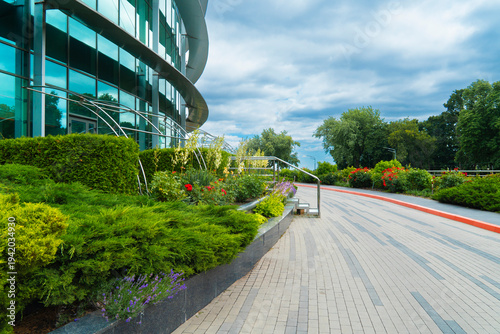 Contemporary business center building with glass facade and professional landscaping featuring green bushes, flowers and a paved pedestrian walkway