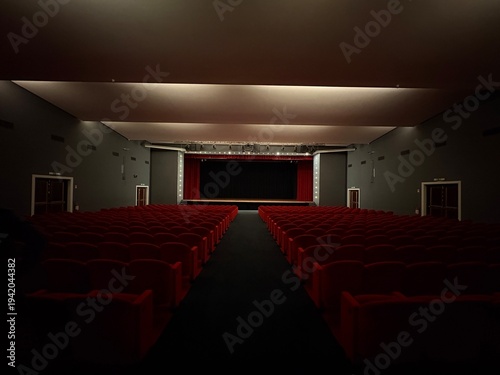 Obraz na plátně Interior of an empty theater auditorium with rows of red seats facing a stage with closed curtain and dramatic lighting