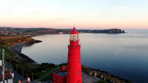 Stunning aerial view of a vibrant red lighthouse by the ocean at sunset.