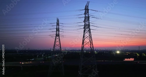 Electricity transmission pylons and high voltage power lines standing against beautiful sunset sky above city highway.