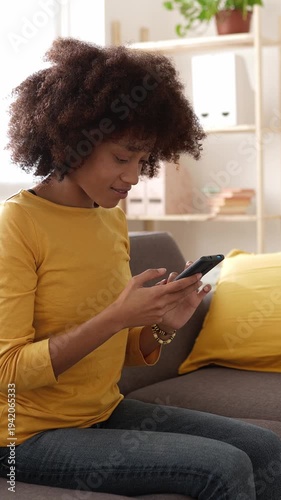 Vertical HD video of young happy black woman sitting on the sofa at home, using her mobile phone, gets excited and celebrates receiving good news, a message, or winning something online