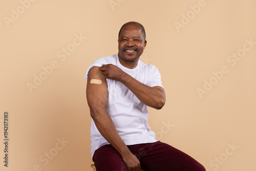 Mature african american man sitting on stool in casual clothing showing bandage on his upper arm on beige studio background. Concept of vaccination, immunization and preventive healthcare